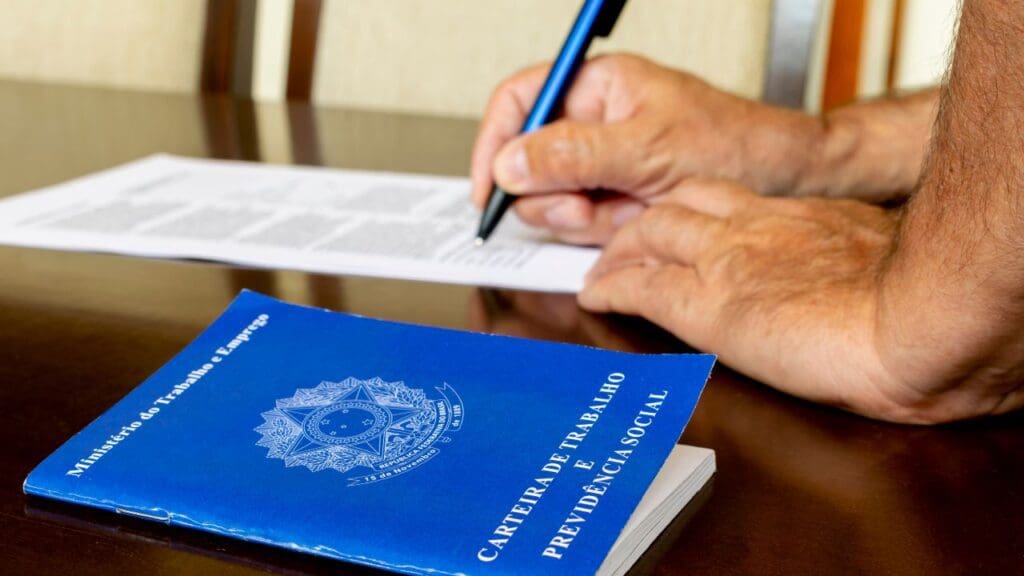 Trabalhador assinando contrato e carteira trabalho ao lado em cima da mesa