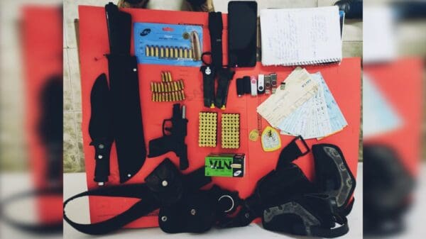 Sobre uma mesa vermelha, arma de fogo, munições, facas, lanterna, pen drives e folhas de cheques