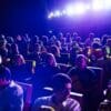 Pessoas sentadas em suas cadeiras em sala de cinema escura com apenas a luz do projetor de filme clareando a sessão