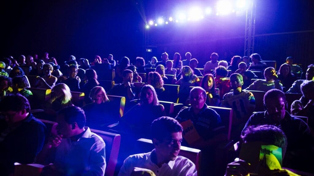 Pessoas sentadas em suas cadeiras em sala de cinema escura com apenas a luz do projetor de filme clareando a sessão