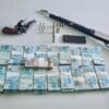 Sobre uma mesa branca da Polícia bolos de notas de 100 reais e duas armas