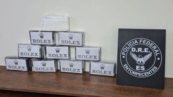 Sobre a mesa da Polícia Federal dez tabletes de cocaína embrulhadas com a marca Rolex, empilhadas e do lado direito uma placa de divulgação da Polícia Federal