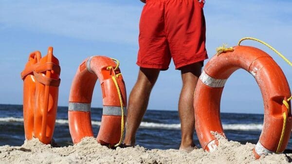 Guarda Vidas parado na praia observando o mar, ao lado dele boias laranjas de resgate
