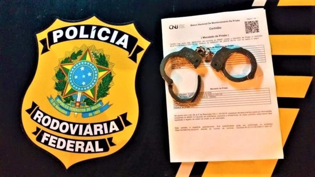 Sobre o Capo da viatura da Polícia Rodoviária Federal um documento de mandado de prisão e uma algema com um dos lados abertos