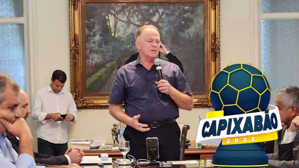 Governador do estado do Espírito Santo Renato Casagrande segurando um microfone e discursando em uma sala de reuniões cheias de dirigentes de futebol e secretários de esportes e na direita da foto o símbolo do Campeonato Capixaba 2023 que é uma bola de futebol azul com detalhes amarelos em cima de uma base azul simulando um troféu com uma faixa branca centralizada escrita Capixabão 2023