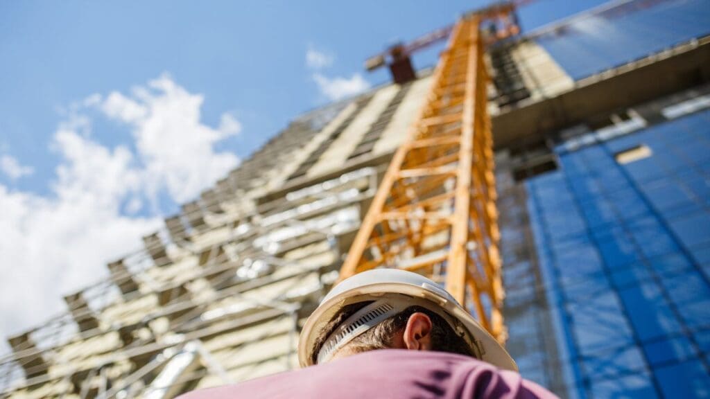 Foto de um funcionário da construção civil de capacete de proteção e ao fundo a imagem desfocada de um edifício enorme em construção