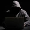 Homem em uma sala escura de moletom cinza e com o capuz na cabeça digitando em um notebook, fazendo referencia a hackers