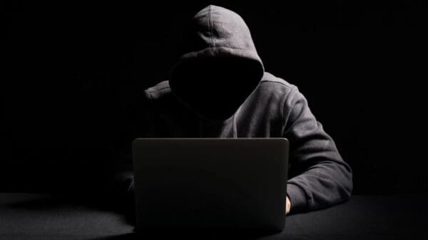 Homem em uma sala escura de moletom cinza e com o capuz na cabeça digitando em um notebook, fazendo referencia a hackers