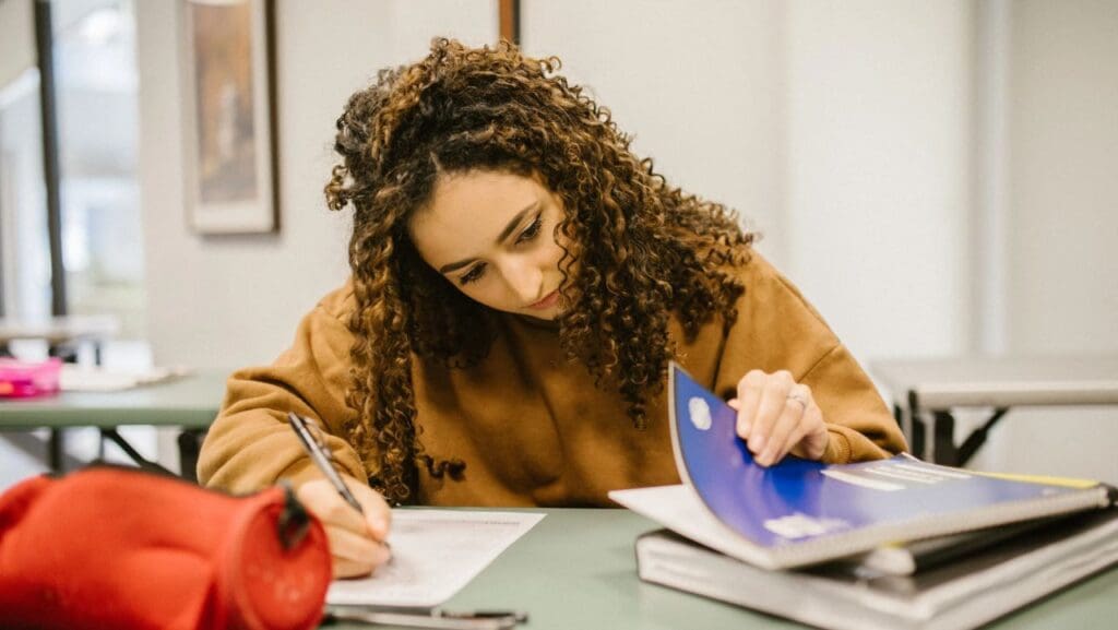 Aluna escrevendo em uma folha com a mão direita e pesquisando um livro com a mão esquerda, na mesa ha um estojo e cadernos