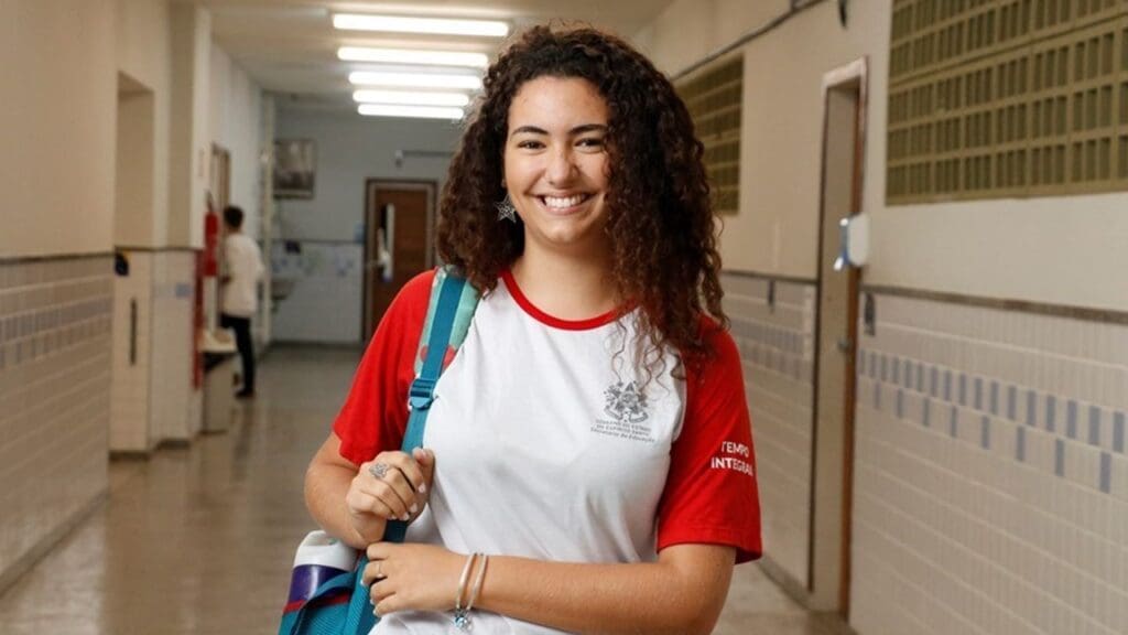 Aluna sorridente segurando a alça da mochila em um corredor de uma escola