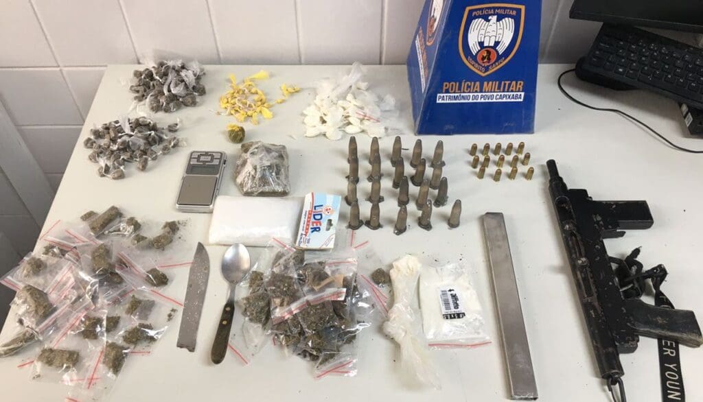 Foto de apreensão de armas e drogas, sobre uma mesa branca uma submetralhadora, um pente, 138 buchas de maconha, uma bola de haxixe, 51 pedras de crack, 48 embrulhos de cocaína e um pedaço de tablete de maconha, além de uma balança de precisão e material para embalo dos entorpecentes