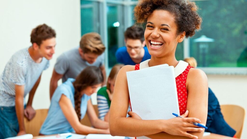 Foto de uma estudante sorridente segurando uma pasta e uma caneta, em destaque e no fundo desfocado 6 estudantes em volta de uma mesa conversando