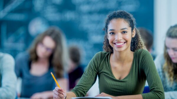 Foto de uma estudante sorridente segurando um lapis enquanto faz uma prova, ao fundo desfocado vemos outros alunos e um lousa