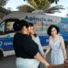 Arte com a Van do Sine móvel em uma praça e em frente a van quatro mulheres sorridentes de mãos dadas