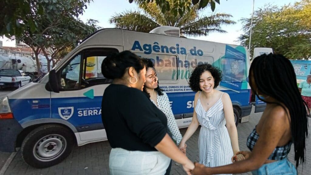 Arte com a Van do Sine móvel em uma praça e em frente a van quatro mulheres sorridentes de mãos dadas