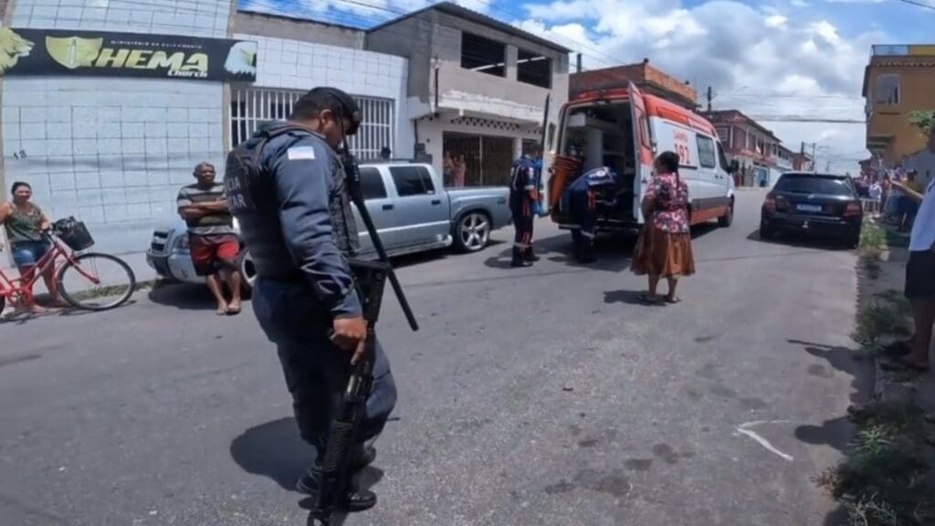 Foto de uma rua do bairro do Jardim Tropical, na foto em destaque vemos um Policial Militar segurando uma segurando escopeta com munição borracha, mais ao fundo uma unidade do SAMU com as portas abertas com um indivíduo que foi contido dentro, ao lada da ambulância um membro da equipe do SAMU e a mãe do indivíduo uma senhora de vestido