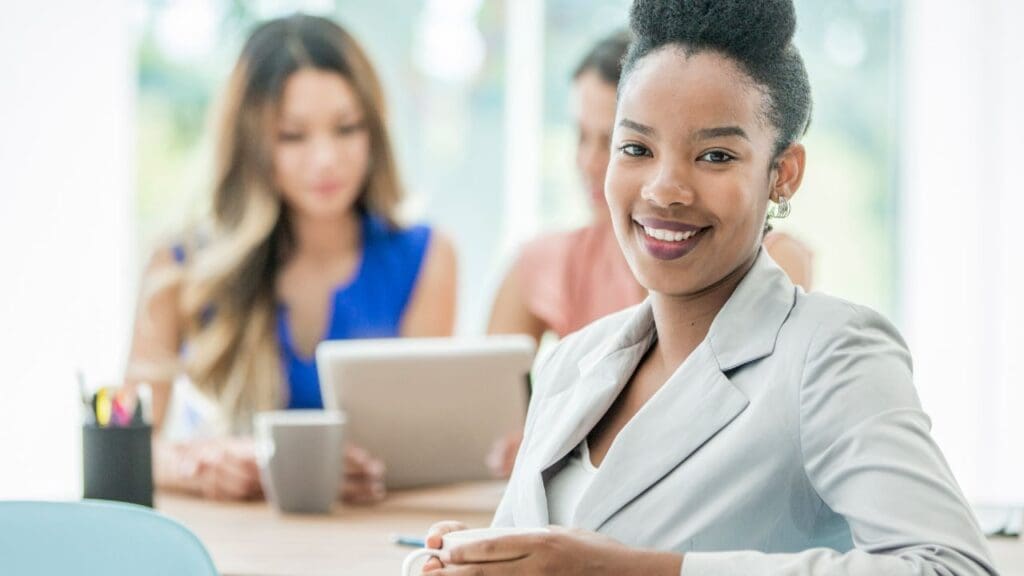 Uma jovem Negra com roupa social sorridente, ao fundo desfocado duas mulheres conversando