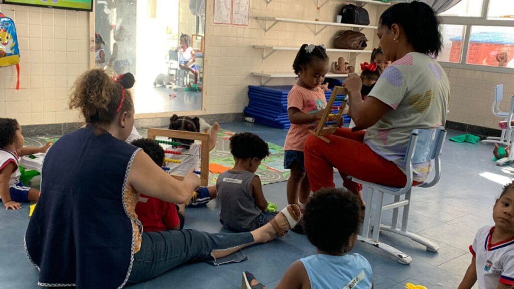 Foto de uma assistente de educação infantil sentada em uma cadeira em uma sala com 7 crianças.