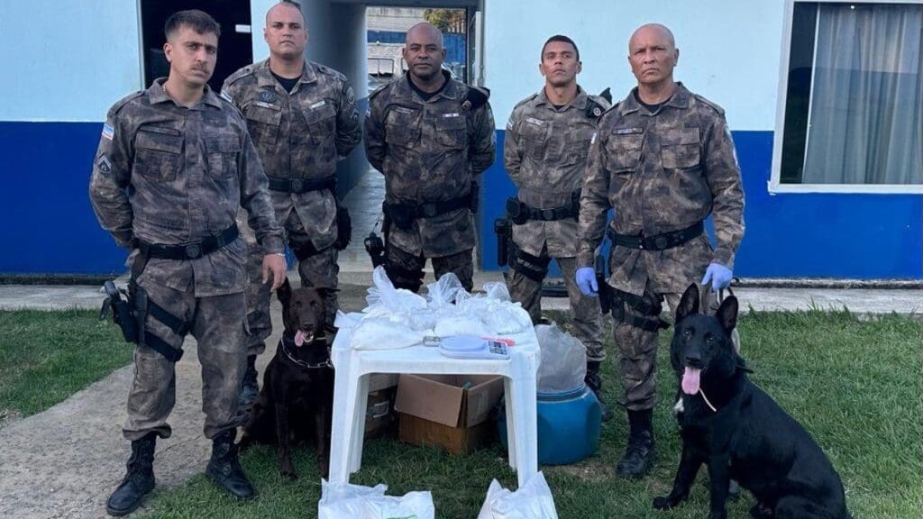 5 membros da Equipe da PM, um deles segurando a coleira de um cachorro pastor alemão preto, no centro da foto uma mesa branca com 43 pedras de crack e duas balanças de precisão, 25 quilos de ácido bórico, 3,8 quilos de adrenalina, mais 3,1 quilos de lidocaína e 600 gramas de anfetamina