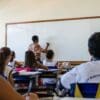 Foto de uma sala de aula com dezenas de crianças estudando enquanto a professora escreve na lousa