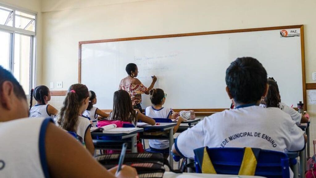 Foto de uma sala de aula com dezenas de crianças estudando enquanto a professora escreve na lousa