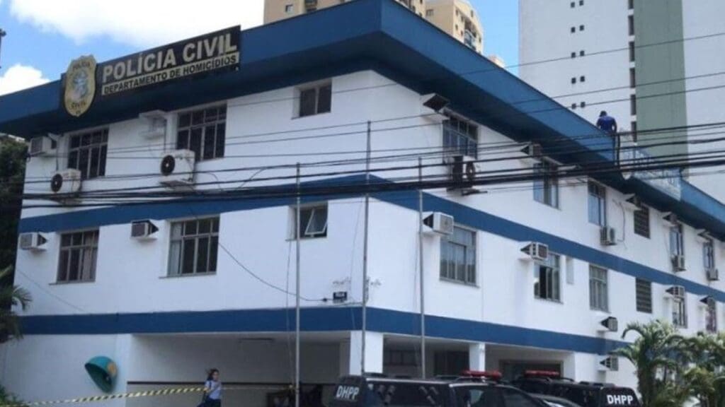 Foto panorâmica da sede Divisão de Homicídios e Proteção à Mulher (DHPM), um prédio de 3 andares, nas cores branca e azul e no patio uma viatura da Polícia Civil