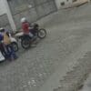 imagem de um veículo branco parado em uma rua de paralelepípedo e uma moto com dois homens assaltando a motorista que está parada em pé ao lado do veiculo