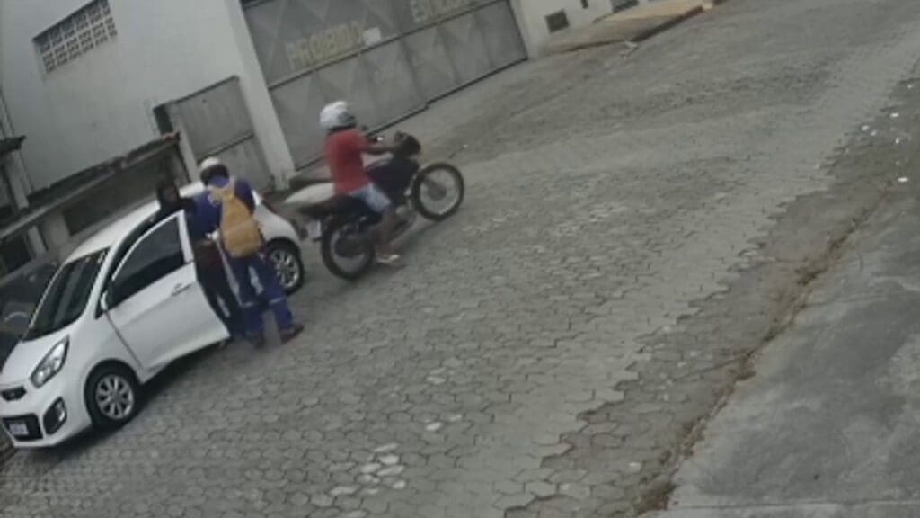 imagem de um veículo branco parado em uma rua de paralelepípedo e uma moto com dois homens assaltando a motorista que está parada em pé ao lado do veiculo