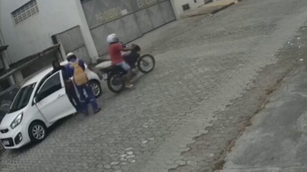 imagem de um veículo branco parado em uma rua de paralelepípedo e uma moto com dois homens assaltando a motorista que está parada em pé ao lado do veiculo