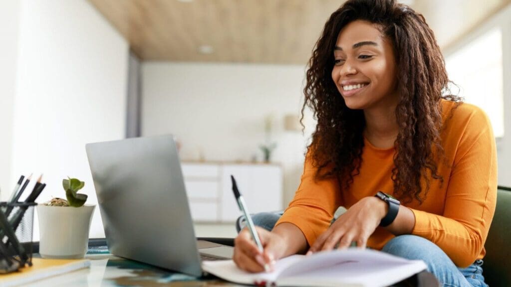 Foto de uma moça estudando no notebook e escrevendo no caderno