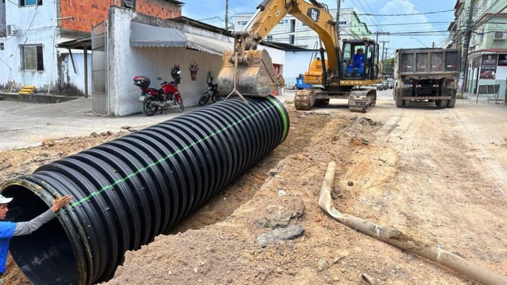 Vias em reforma onde uma máquina está colocando grande tubo enquanto um funcionário homem está apoiando.