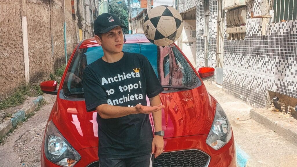 Gabriel Siqueira, idealizador do Anchieta na Alta em frente a um veículo vermelho jogando uma bola de futebol para cima