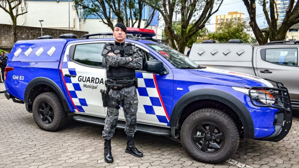 Uma viatura da Guarda Civil Municipal de Vitória, estacionada em um patio com um agente da GCMV em frente com os braços cruzados posando para a foto