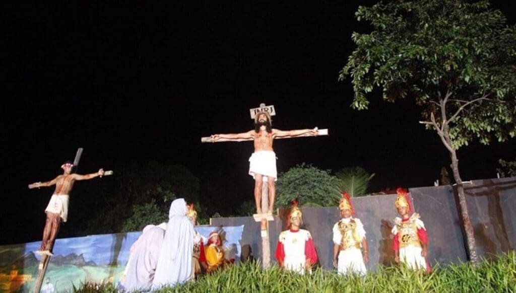 Foto da encenação da Paixão de Cristo, duas pessoas sendo crucificadas uma delas representa Jesus e envolta da crus em que Jesus está soldados romanos e seguidores