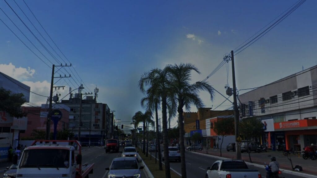 Foto da Avenida Fernando Ferrari uma importante avenida de Vitória que corta vários bairros na imagem dos dois lados da avenida há comércios e agencias de banco, o canteiro central tem palmeiras e a rua está com movimento alto de veículos.