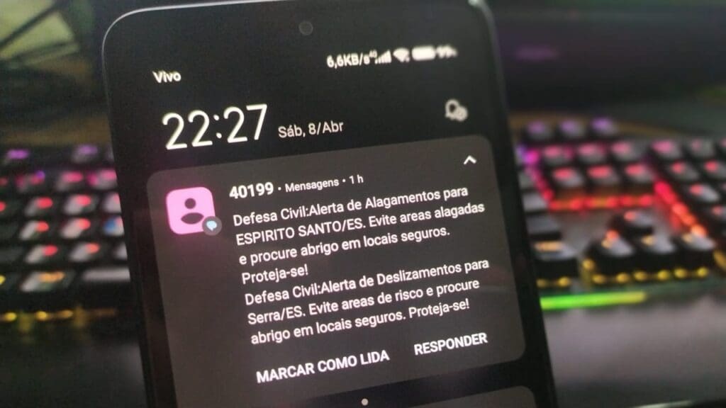 Foto de um celular com dois avisos da defesa civil enviados por SMS, no primeiro: Defesa Civil: Alerta de Deslizamentos para Serra/ES. Evite áreas de risco e procure abrigo em locais seguros. Proteja-se! e na segunda mensagem: Defesa Civil: Alerta de Deslizamentos para Serra/ES. Evite áreas de risco e procure abrigo em locais seguros. Proteja-se! e no fundo desfocado é possivel ver um teclado gamer com leds acesos