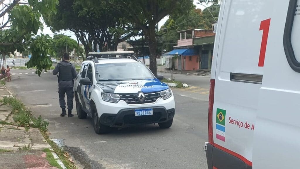 Foto uma viatura da Polícia Militar parada em uma rua, bem arborizada com um soldado parado de costas ao lado esquerdo da viatura, e alguns metros a frente é possível ver uma unidade do SAMU