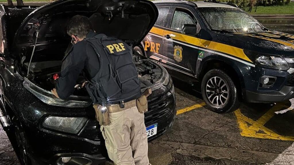 Foto de um agente da PRF vistoriando um veículo Fiat Toro, que está com o capo aberto ao lado uma viatura da PRF