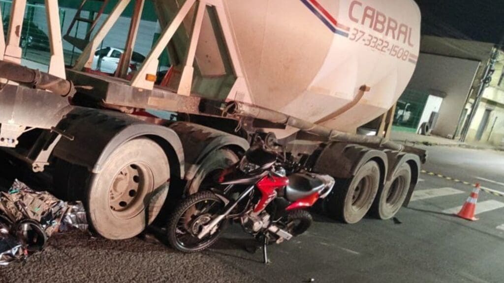 Foto com uma carreta estacionada ao lado de uma Moto Honda/XRE 300 vermelha com o pneu traseiro amassado após acidente fatal