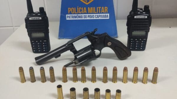 Sobre uma mesa branca, um revólver calibre 38 com a numeração raspada, cinco munições intactas e uma deflagrada, e dois rádio comunicador