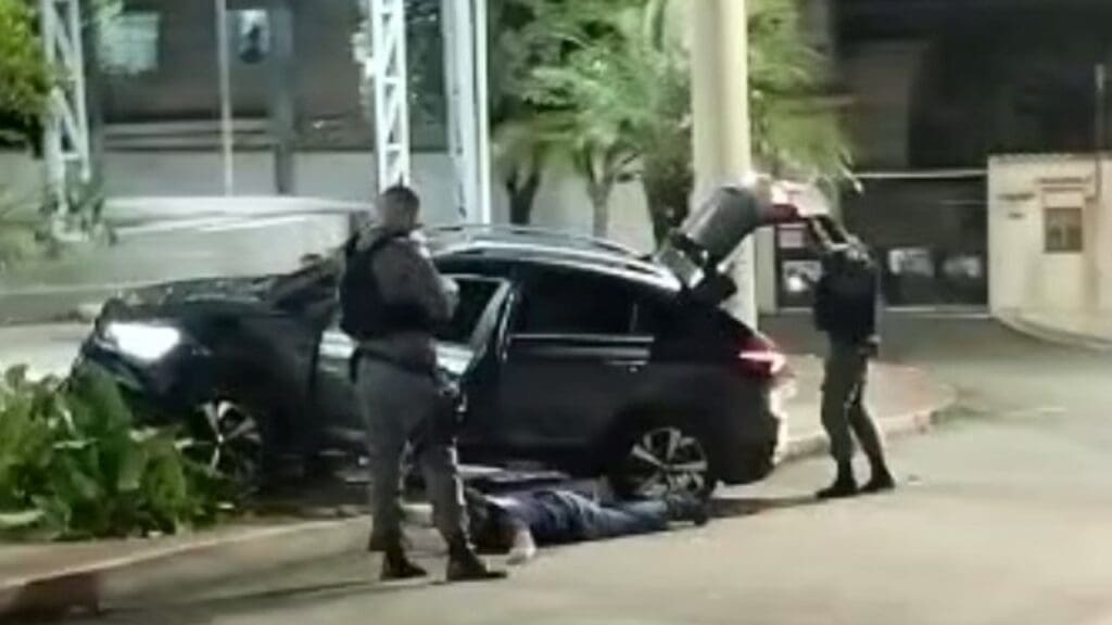 Foto dois Policiais Militares revistando um veículo, enquanto o motorista está no chão aguardando a revista