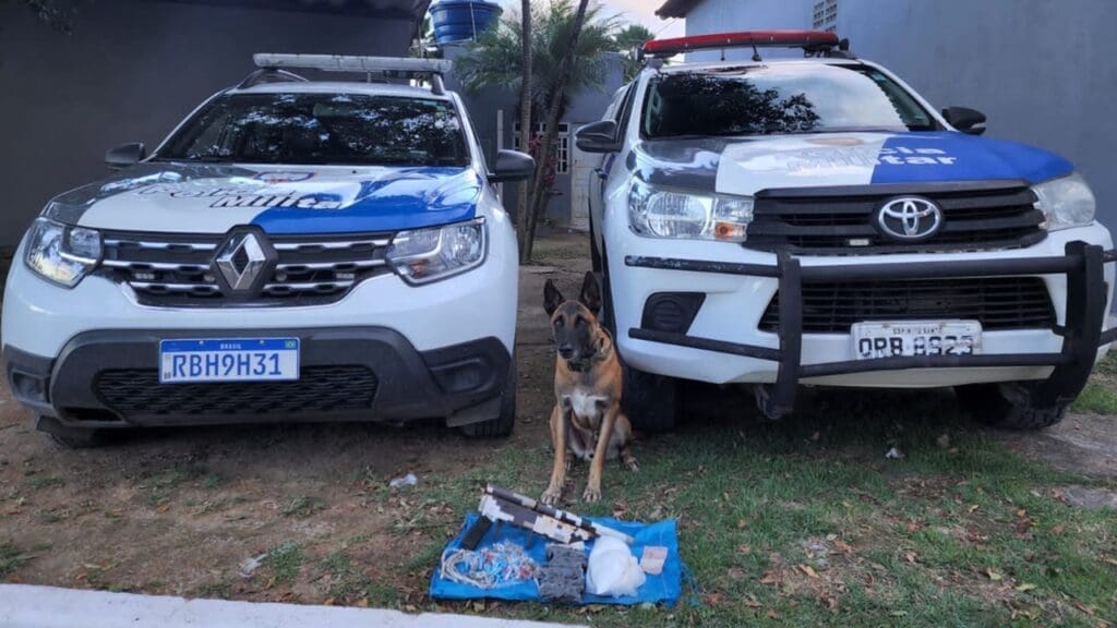 Foto do cão Apollo da PM, parado e em sua frente materiais apreendidos 01 escopeta calibre 12, de fabricação semi industrial, 01 munições calibre 12, 089 buchas de maconha (130g), 073 pedras de crack, 275 pinos de cocaína (425g), 01 porção de cocaína (905g), 05 reais em espécie e duas viaturas ao fundo.