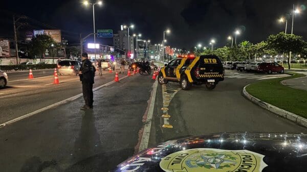 Uma avenida durante a noite com viaturas da PRF e agentes parando veículos, durante uma blits