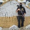Um paredão de com centenas de tabletes de drogas e um Agente da Polícia Militar armado e de capuz posando para a foto de divulgação.