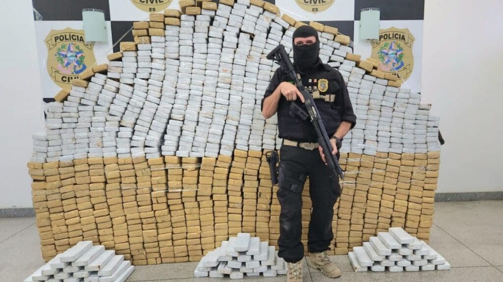 Um paredão de com centenas de tabletes de drogas e um Agente da Polícia Militar armado e de capuz posando para a foto de divulgação.