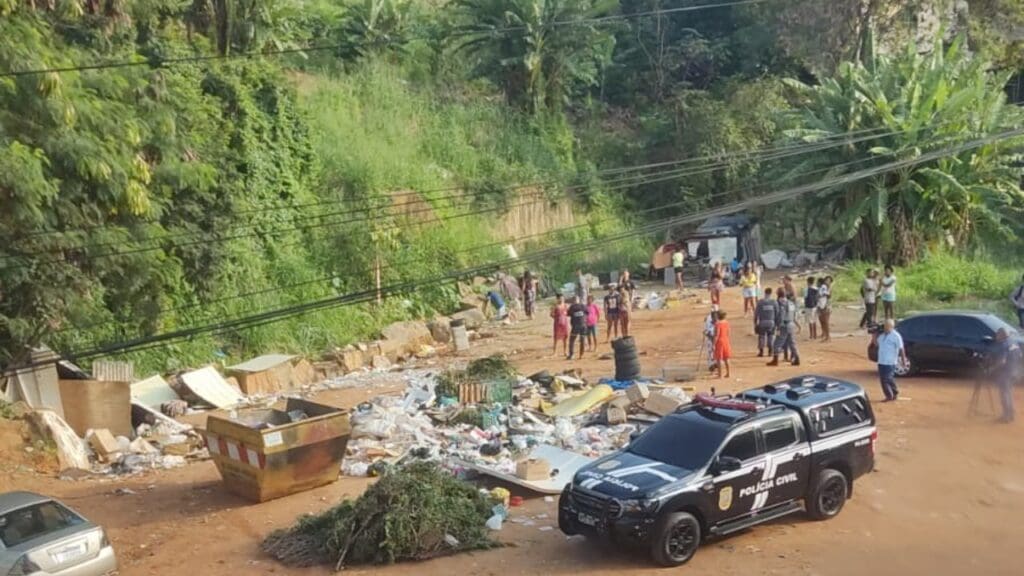 Foto de um terreno abandonado, onde foi encontrado um corpo de um homem de 33 anos assassinado a tiros , no local é possível ver uma viatura da Polícia Civil e muitos moradores do local em volta do corpo