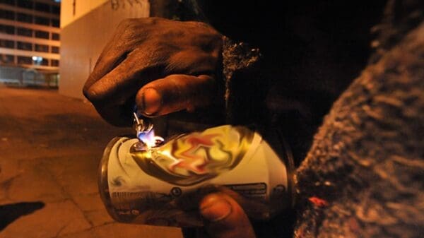 Homem usando uma lata para fumar crack em um local escuro com predios em volta