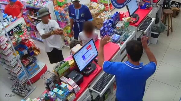 Foto do momento que homens armados anunciam o assalto a uma farmácia, do outro lado do balcão um funcionário, com as mãos para cima.