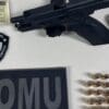 Foto sobre uma mesa branca, uma pistola 9mm, com um pente preso no local do gatilho, uma carteira de motorista falsa, 22 munições da pistola e 2 flamulas da Guarda da Serra/ROMU.