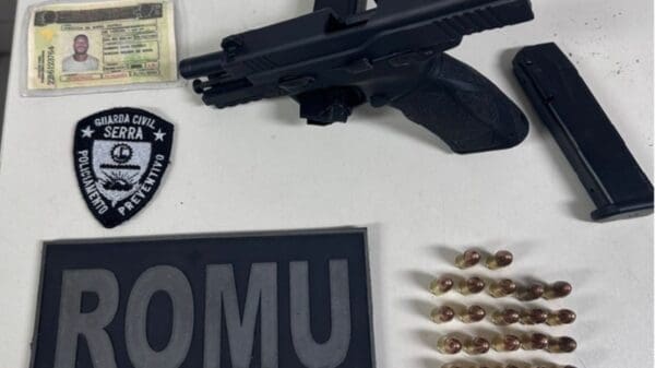 Foto sobre uma mesa branca, uma pistola 9mm, com um pente preso no local do gatilho, uma carteira de motorista falsa, 22 munições da pistola e 2 flamulas da Guarda da Serra/ROMU.